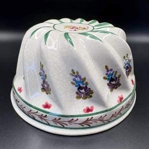 May include: White ceramic bundt pan with a floral design. The pan features a crackle glaze and is decorated with green leaves, red and purple flowers, and a green border. The pan has a scalloped edge.