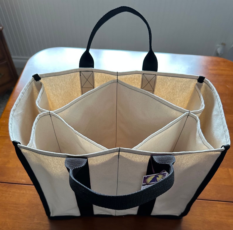 May include: A natural canvas tote bag with black handles and black trim. The bag has seven separate compartments, each with a black stitched border.