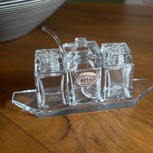 Könnte beinhalten: Ein durchsichtiges Glas-Gewürzset mit drei quadratischen Behältern und einem Löffel, das auf einem rechteckigen Tablett ruht. Das Set enthält einen zentralen Behälter mit Deckel und zwei kleinere Behälter mit Schraubdeckeln.