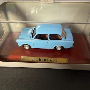 May include: A light blue Trabant 601 model car displayed in a clear case. The miniature car has a brown interior and silver wheels. The model sits on a wooden base with a gold nameplate.