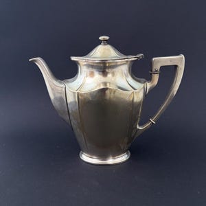 Könnte beinhalten: Eine silberfarbene Teekanne mit Deckel und Griff. Die Teekanne hat einen gebogenen Ausguss und einen quadratischen Griff. Der Körper der Teekanne hat ein geriffeltes Design. Die Teekanne steht vor einem dunklen Hintergrund.