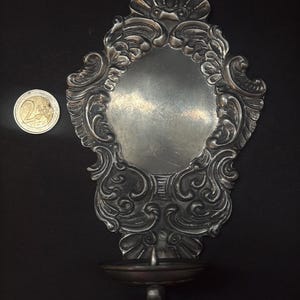 May include: A silver-toned metal wall sconce featuring an ornate, embossed scrollwork backplate with a shell motif at the top and a circular candle holder at the base. A 2 euro coin is placed to the left for scale, showing the relative size.