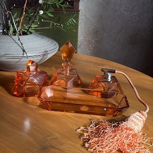 Könnte beinhalten: Ein orangefarbenes Parfümflaschen-Set aus Glas mit geometrischen Formen, darunter eine Flasche mit Kristallverschluss, eine Ablage und zwei kleinere Flaschen. Eine dekorative Quaste befindet sich neben dem Set. Das Set steht auf einem Holztisch.