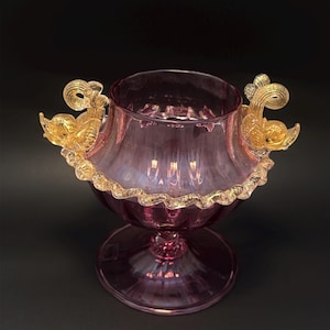 May include: A pink glass bowl with gold accents. The bowl has a ruffled gold rim and two gold swan-shaped handles. The bowl sits on a pedestal base, and the background is black.