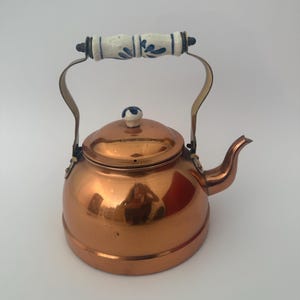 Könnte beinhalten: Ein kupferfarbener Teekessel mit einem weiß-blauen floral gemusterten Griff. Der Kessel hat einen abgerundeten Körper, einen Ausguss und einen Deckel mit einem kleinen Knopf. Der Griff ist gebogen und aus messingfarbenem Metall.