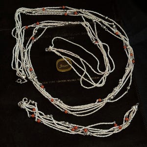 Könnte beinhalten: Ein mehrsträngiges Set aus Silberhalskette und Armband mit feinen, gedrehten Silberketten, die mit kleinen, runden, orange-roten Korallenperlen besetzt sind. Die Schmuckstücke sind kreisförmig auf einer dunkelbraunen Stoffoberfläche angeordnet.