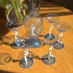 Könnte beinhalten: Ein Set aus eleganten Gläsern mit einer blauen Glaskaraffe und sechs passenden Stielgläsern. Jedes Glas wird von einer weißen Figur getragen. Das Glaswaren-Set steht auf einer Holzoberfläche, mit einer Vase mit grünen Blättern im Hintergrund.