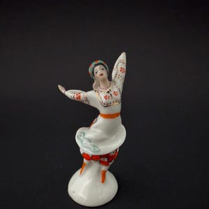 Könnte beinhalten: Porzellanfigur einer tanzenden Frau in traditioneller Kleidung. Die Figur trägt ein weißes Kleid mit roten Blumenmustern, einen orangefarbenen Gürtel und ein gemustertes Kopftuch. Die Figur steht auf einem weißen Sockel und hat die Arme ausgestreckt.