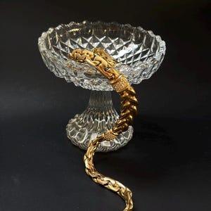 Könnte beinhalten: Ein goldfarbenes Armband mit geflochtenem Design, verziert mit kleinen, funkelnden Verzierungen, liegt in einer transparenten, geschliffenen Glasschale auf einem Sockel. Die Schale hat ein Rautenmuster und einen runden Fuß. Der Hintergrund ist schwarz.