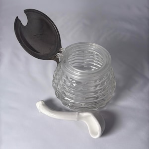 May include: A clear glass jar with a textured, ridged pattern, topped with a hinged, dark metal lid. A white ceramic spoon rests beside the jar. The jar and spoon are on a white surface.