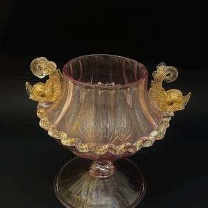 May include: A decorative glass vase with a flared rim and a rounded base. The vase is a soft pink color with gold accents and two ornate gold handles shaped like mythical creatures. The rim is trimmed with a gold, textured border.