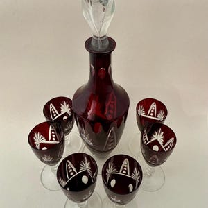 Könnte beinhalten: Ein Satz rubinroter Glaswaren, bestehend aus einer Karaffe mit einem durchsichtigen Verschluss und sechs Weingläsern. Jedes Stück hat geätzte weiße Muster. Ein klassisches Vintage-Glaswarenset.