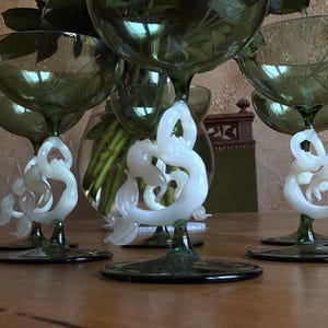 Könnte beinhalten: Eine Sammlung grüner Sektgläser mit weißen, wirbelnden, skulpturalen Stielen. Die Gläser stehen auf einem Holztisch, im Hintergrund eine Vase mit Blumen. Die Gläser haben ein Blattmuster, das in die Schale geätzt ist.