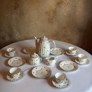Może przedstawiać: Biały zestaw do herbaty z porcelany o żebrowanym wzorze i delikatnym kwiatowym motywie z niebieskim wykończeniem. Zestaw zawiera wysoki czajnik, cukiernicę, dzbanek na mleko, cztery filiżanki i cztery pasujące spodki, ustawione na białej powierzchni.