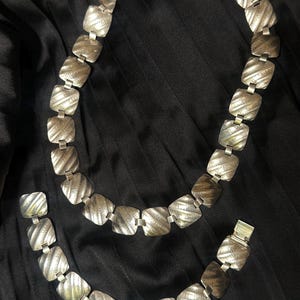 Könnte beinhalten: Ein silberfarbenes Halsketten- und Armband-Set. Der Schmuck weist ein sich wiederholendes Muster aus quadratischen, strukturierten Gliedern auf. Das Set wird auf einem schwarzen, plissierten Stoffhintergrund präsentiert.