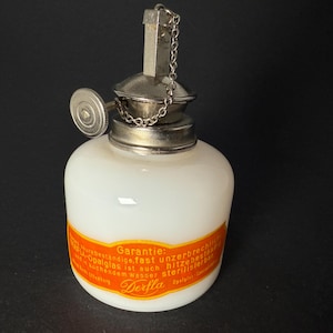 May include: A white glass Derfla lamp with a metal top and chain. The lamp has a round body and a metal cap with a small chain attached. The lamp has a red and yellow label with German text.
