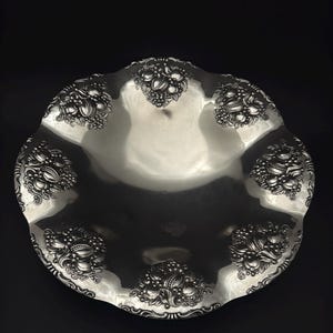 Könnte beinhalten: Eine silberfarbene, verzierte Servierschale mit gewelltem Rand. Die Schale weist detaillierte Blumen- und Fruchtdesigns am Rand auf. Der Innenraum ist glatt und reflektierend, vor dunklem Hintergrund. Ein dekoratives Stück zum Servieren von Speisen.