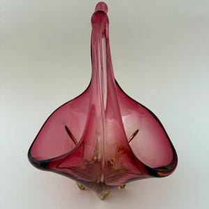 Könnte beinhalten: Eine antike Glasschüssel für Obst mit einem einzigartigen, abstrakten Design. Die Schüssel besteht aus klarem Glas mit einem rötlich-violetten Farbton und einem goldfarbenen Boden. Die Schüssel hat einen einzigen Griff, der aus der Mitte der Schüssel herausragt.