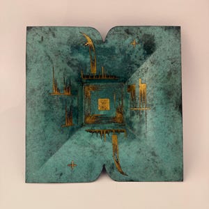May include: Square, teal-colored decorative object with gold accents. The object features a central square with a smaller gold square inside, surrounded by gold geometric shapes and lines. The edges have a unique, curved design.