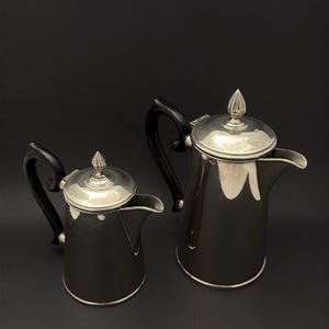 Könnte beinhalten: Zwei silberfarbene Kaffeekannen mit schwarzen Griffen und Deckeln. Die Kannen sind unterschiedlich groß, eine kleinere links und eine größere rechts. Die Deckel haben einen dekorativen Knauf. Die Kannen sind wahrscheinlich aus Metall.