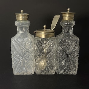 Könnte beinhalten: Drei klare Glasflaschen mit silberfarbenen Deckeln. Die Flaschen haben ein rautenförmiges Design. Die mittlere Flasche hat einen Klappdeckel und einen kleinen Löffel. Die Flaschen dienen wahrscheinlich zum Servieren von Gewürzen.