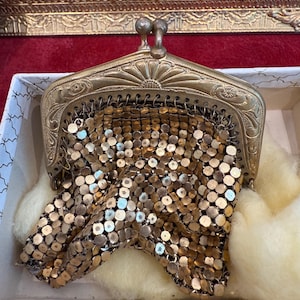 Könnte beinhalten: Eine goldfarbene Mesh-Handtasche mit einem dekorativen Metallrahmen. Die Tasche ist geschlossen und befindet sich in einer weißen Schachtel mit roter Samtfütterung.