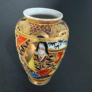 Puede incluir: Un pequeño jarrón japonés decorativo con interior blanco y borde dorado. El jarrón presenta un diseño detallado con una mujer con vestimenta tradicional, sobre un fondo de patrones rojos, negros y dorados. El jarrón mide aproximadamente 10 cm de alto.