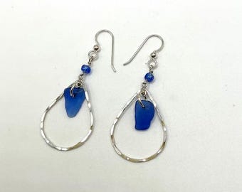 May include: A pair of drop earrings featuring silver-toned hammered teardrop hoops. Each earring is accented with a small, vibrant blue glass bead and a single, irregular piece of cobalt blue sea glass suspended from the center of the hoop.
