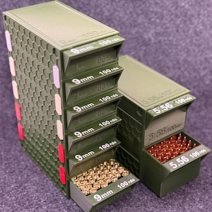 Könnte beinhalten: Olivgrüne Munitionsaufbewahrungsbehälter mit ausziehbaren Schubladen. Die Schubladen sind mit "9mm 100 rds" und "5.56 100 rds" beschriftet. Die Behälter sind gestapelt und haben rote, weiße und beige Akzente. Das Bild zeigt auch lose Teile.