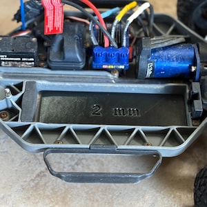 Può includere: Primo piano del telaio grigio di un'auto telecomandata. Il telaio ha una maniglia nera e un vano con la scritta "2 mm". I componenti visibili includono un motore blu, fili e un connettore blu.