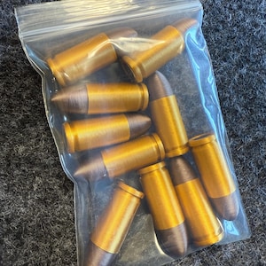 May include: A clear plastic bag filled with several metallic gold-colored bullets with brown tips. The bullets are cylindrical with a ridged texture and are arranged in a haphazard manner within the bag.