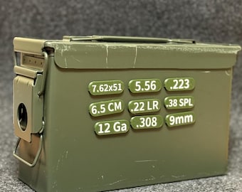 May include: An olive green metal ammunition box with a hinged lid and a latch. The box has several labels with text including "7.62x51", "5.56", ".223", "6.5 CM", ".22 LR", ".38 SPL", "12 Ga", ".308", and "9mm".