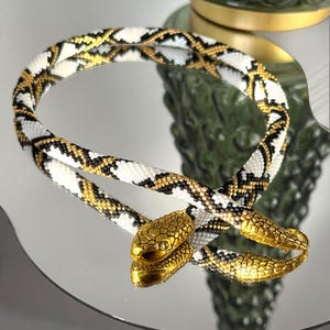Puede incluir: Un collar de serpiente de cuentas blancas, negras y doradas con detalles de cabeza de serpiente dorada.