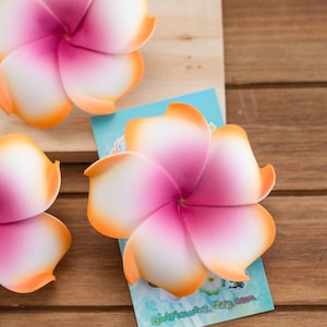 Könnte beinhalten: Drei rosa und orangefarbene Frangipani-Blüten mit weißen Zentren. Die Blüten sind aus Schaumstoff und werden als Haarschmuck verwendet.