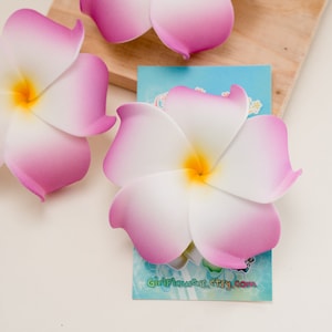 May include: Three pink and white plumeria flowers with yellow centres. The flowers are made of foam and are used for hair accessories or decorations. The flowers are on a wooden surface with a blue card that says "GirlFlowers.etsy.com".