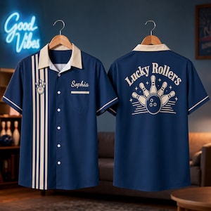 May include: A navy blue short-sleeve button-down shirt with a cream-coloured collar and vertical stripes on the front. The front features a small bowling pin graphic and the name Sophia. The back displays a bowling graphic with the text Lucky Rollers.