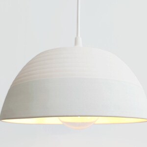 May include: A white ceramic pendant light with a textured, ribbed top and a smooth bottom. The light is hanging from a white cord.