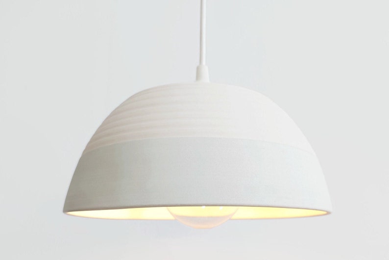 May include: A white ceramic pendant light with a textured, ribbed top and a smooth bottom. The light is hanging from a white cord.