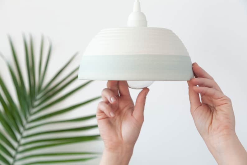 May include: A white ceramic pendant light with a light blue band around the bottom. The light is being installed by a person.