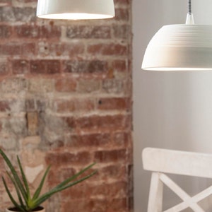 May include: Two white ceramic pendant lights hanging from the ceiling against a brick wall background. The lights are both on and casting a warm glow.