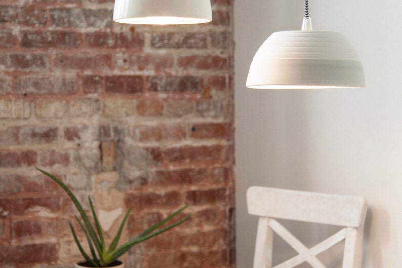 May include: Two white ceramic pendant lights hanging from the ceiling against a brick wall background. The lights are both on and casting a warm glow.