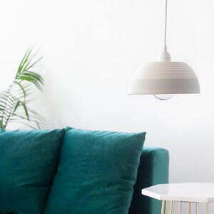 May include: A teal velvet sofa with three pillows sits in a room with a white pendant light hanging from the ceiling. A white side table with a gold metal base sits in front of the sofa.
