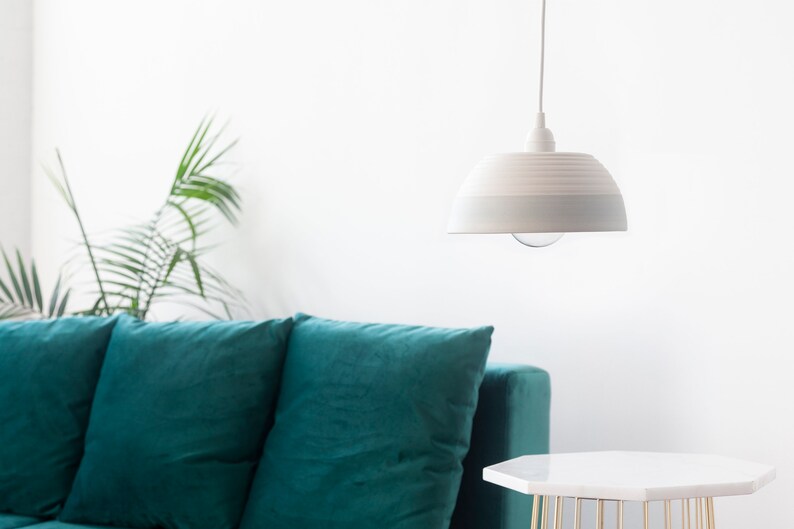 May include: A teal velvet sofa with three pillows sits in a room with a white pendant light hanging from the ceiling. A white side table with a gold metal base sits in front of the sofa.