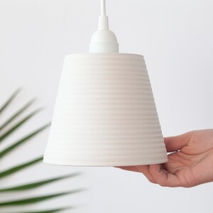 May include: White ceramic pendant light with a ribbed design. The light is hanging from a white cord.