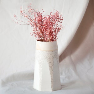 Puede incluir: Un jarrón de cerámica blanco con textura moteada y diseño geométrico. El jarrón está lleno de flores secas rosas.