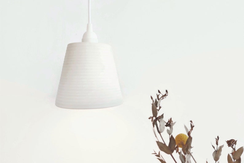 May include: A white ceramic pendant light with a ribbed texture hangs from a white ceiling. The light is off and the background is a white wall.
