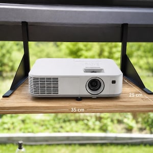 Op de afbeelding: Een witte digitale projector staat op een rechthoekige houten plank die wordt ondersteund door twee zwarte metalen beugels. De plank is 35 cm breed en 25 cm diep. De projector is voorzien van een lens aan de voorzijde en een ventilatierooster.