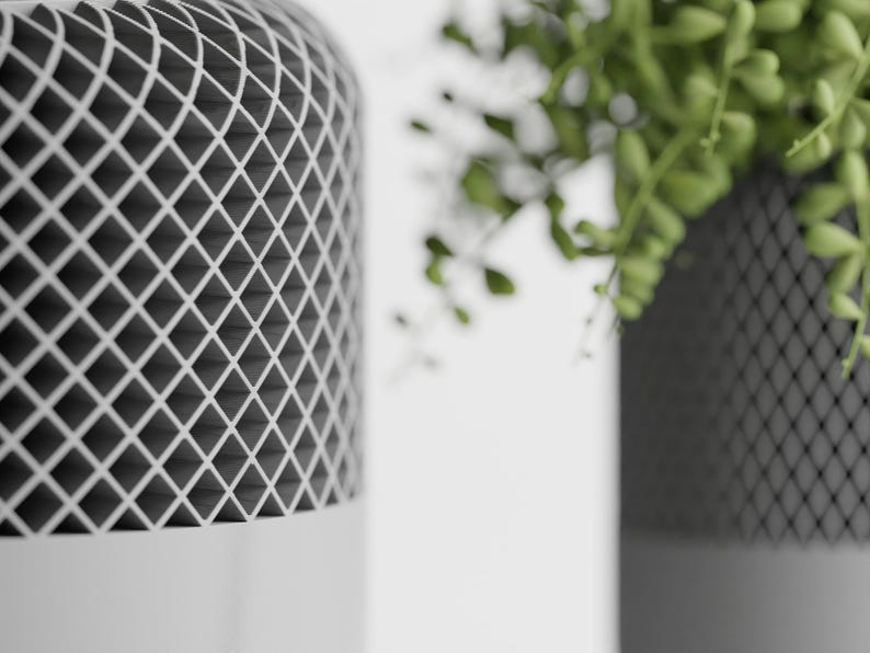 May include: Two white cylindrical planters with a black grid pattern. The planter on the right has a green plant growing out of it.