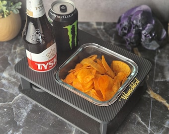 May include: A black serving tray with a carbon fiber pattern holds a bottle of beer, a can of energy drink, and a glass dish filled with chips. The tray has the word "Wunschtext" in gold script.