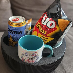 May include: A black, round, multi-compartment tray holds a can of Rockstar energy drink, a bag of Tio Nacho cheese tortilla chips, and a white mug with a blue handle and interior. The mug has floral and text designs.